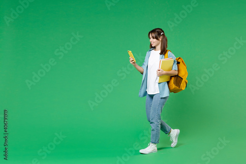 Full length side view of woman student in shirt backpack hold notebooks using mobile cell phone typing sms message isolated on green background. Education in high school university college concept.