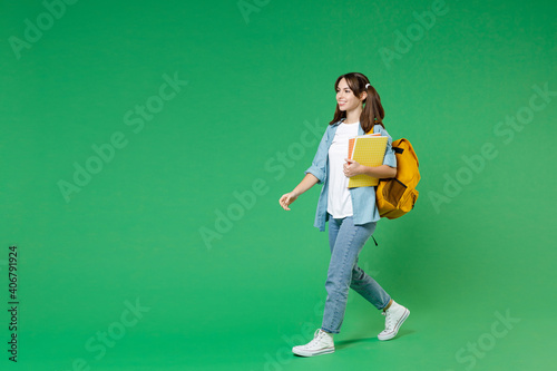 Full length side view of funny young woman student in blue shirt backpack hold notebooks walking going isolated on green color background studio. Education in high school university college concept.