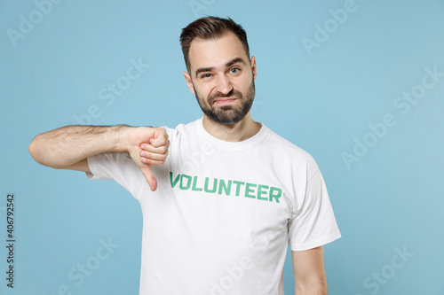 Displeased confused young bearded man in white volunteer t-shirt showing thumb down looking camera isolated on blue background studio portrait. Voluntary free work assistance help charity concept. photo