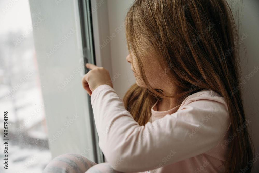 Little girl in pink pajamas sits near the window. Quarantine. Lockdown.