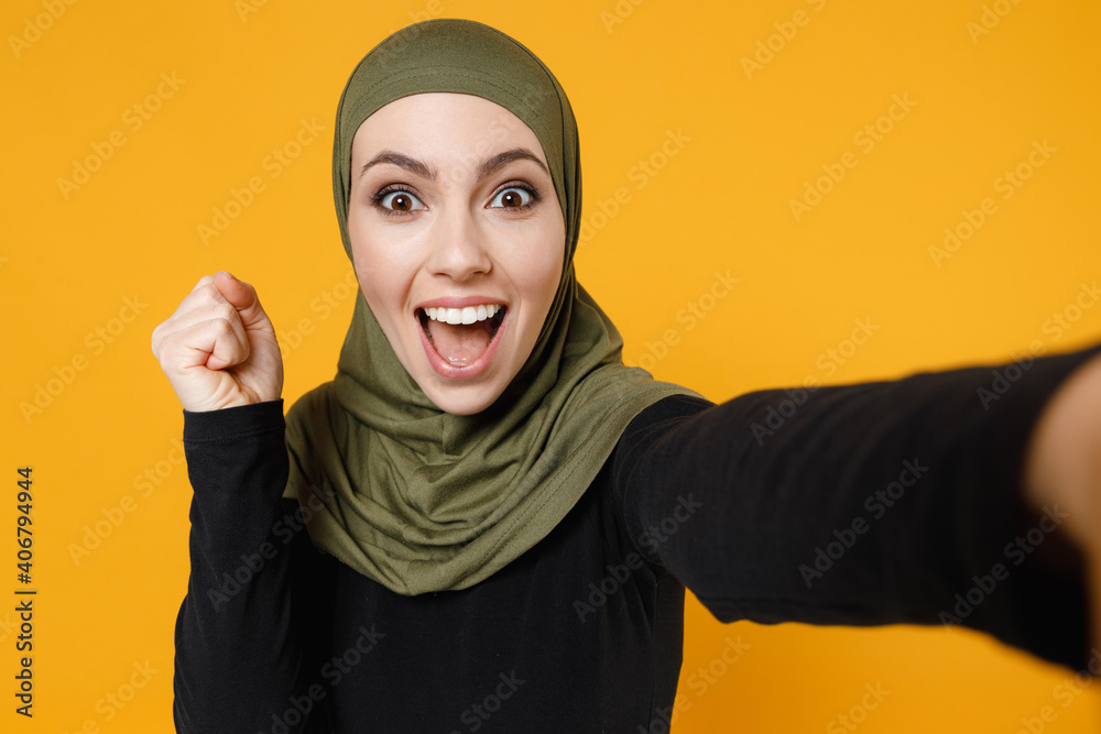 Close up excited young arabian muslim woman in hijab black green clothes doing selfie shot on mobile phone clenching fist like winner isolated on yellow background. People religious lifestyle concept. Close up excited young arabian muslim woman in hijab black green clothes doing selfie shot on mobile phone clenching fist like winner isolated on yellow background. People religious lifestyle concept.