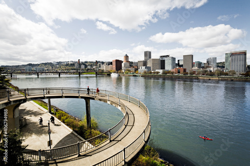 Looking over the Eastbank Esplanade towards downtown Portland on a sunny day.  A handful of people are walking in the foreground and a kayaker rows by in the river.