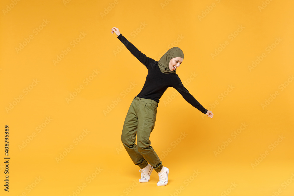 Full length of excited young arabian muslim woman in hijab black green clothes dancing stand on toes spreading hands isolated on yellow background studio portrait. People religious lifestyle concept.