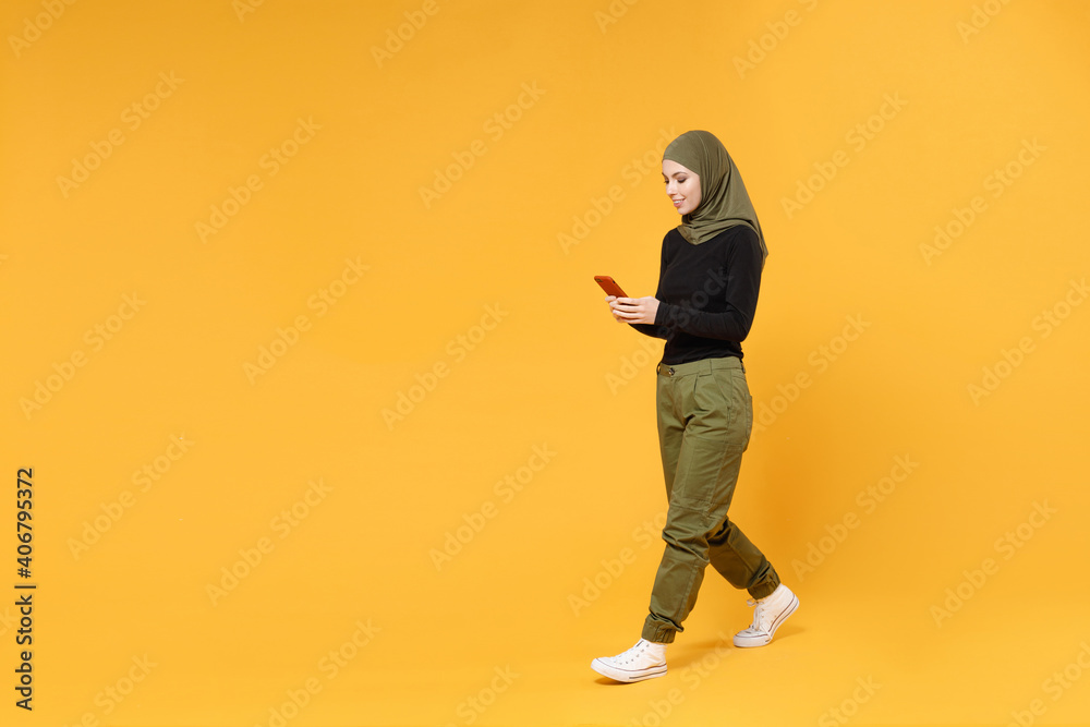 Full length side view of smiling young arabian muslim woman in hijab black green clothes using mobile cell phone typing sms message isolated on yellow background. People religious lifestyle concept. Full length side view of smiling young arabian muslim woman in hijab black green clothes using mobile cell phone typing sms message isolated on yellow background. People religious lifestyle concept.