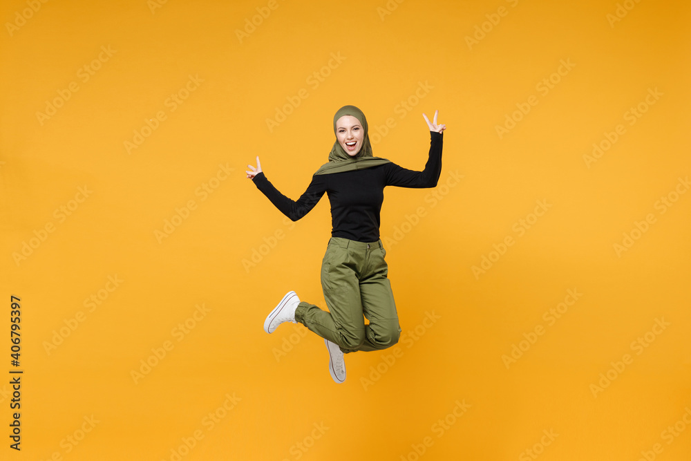 Full length excited young arabian muslim woman wearing hijab black green clothes jumping showing victory sign isolated on yellow color background studio portrait. People religious lifestyle concept.