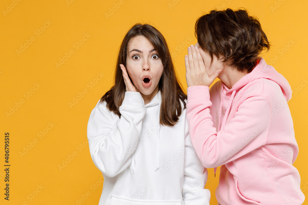 Shocked amazed two young women friends 20s in casual white pink hoodies whispering gossip and tells secret behind her hand, sharing news isolated on bright yellow color background studio portrait.