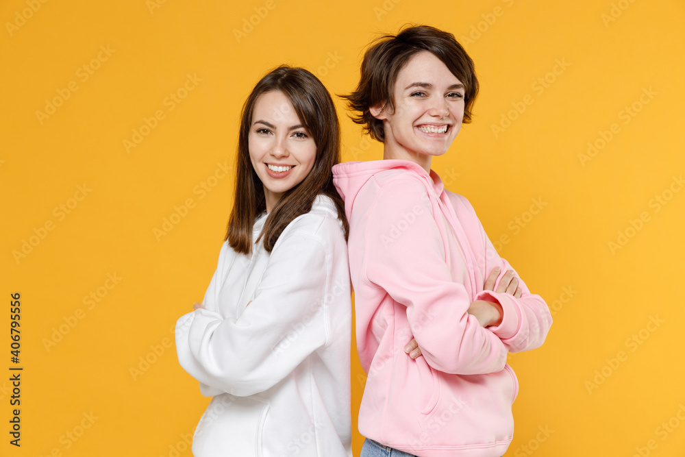 Smiling attractive two young women friends 20s wearing casual white pink hoodies standing back to back holding hands crossed looking camera isolated on bright yellow color background studio portrait. Smiling attractive two young women friends 20s wearing casual white pink hoodies standing back to back holding hands crossed looking camera isolated on bright yellow color background studio portrait.