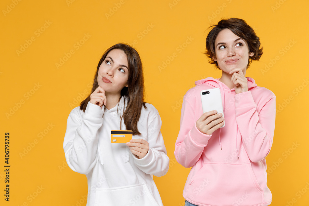Pensive two young women friends 20s wearing casual white pink hoodies standing using mobile cell phone hold credit bank card put hand prop up on chin isolated on yellow background studio portrait. Pensive two young women friends 20s wearing casual white pink hoodies standing using mobile cell phone hold credit bank card put hand prop up on chin isolated on yellow background studio portrait.