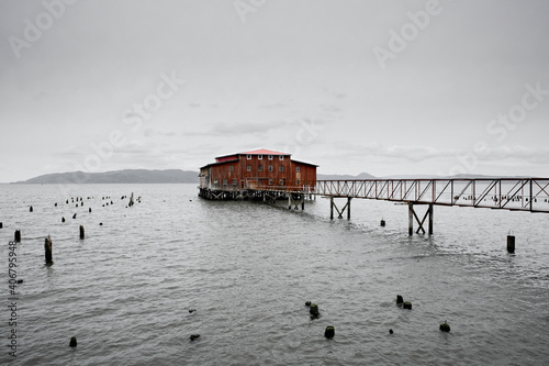 An old red building sits at the end of a wooden dock in the middle of gray body of water.  It's an overcast and gloomy day.  photo