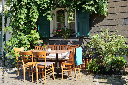 Close-up of a cute table with a bench and chairs in front of a slate plated house with green shutters and decorated with plants