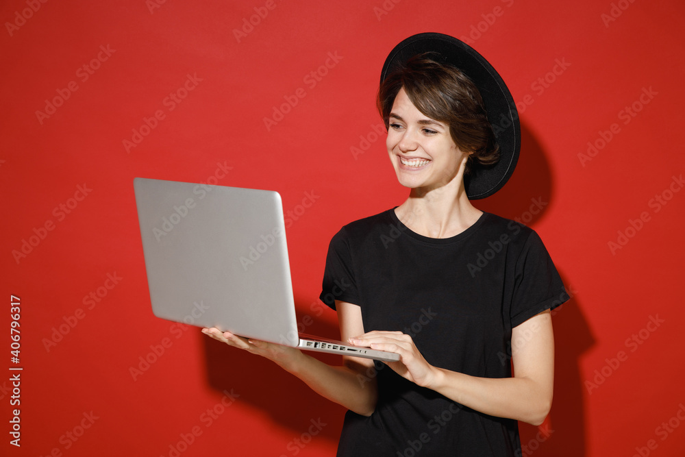 Smiling cheerful beautiful young brunette woman 20s years old wearing casual basic black t-shirt hat standing working on laptop pc computer isolated on bright red color background studio portrait. Smiling cheerful beautiful young brunette woman 20s years old wearing casual basic black t-shirt hat standing working on laptop pc computer isolated on bright red color background studio portrait.