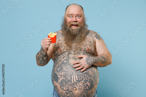 Excited fat pudge obese chubby overweight man has tattooed naked bare big belly hold french fries potatoes put hand on stomach isolated on blue background. Weight loss obesity unhealthy diet concept.