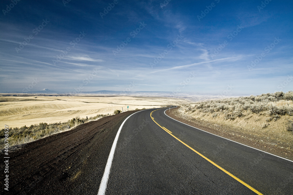 A single lane highway curves around a very low hill in the high desert of Eastern Oregon. A dry, arid landscape stretches on for miles in the distance. A single lane highway curves around a very low hill in the high desert of Eastern Oregon. A dry, arid landscape stretches on for miles in the distance.