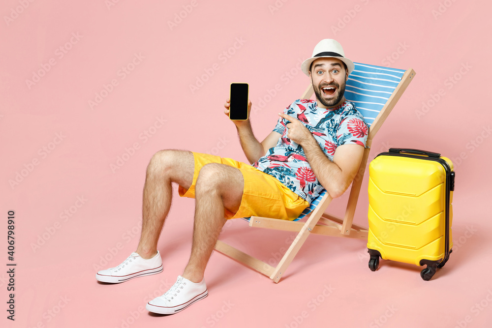 Full length amazed tourist man in hat sit on deck chair point index finger on mobile phone with blank empty screen isolated on pink background. Passenger travel on weekend. Air flight journey concept. - obrazy, fototapety, plakaty 