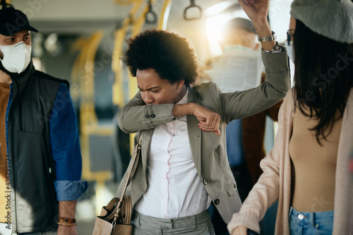 Black businesswoman sneezing into elbow while commuting to work by bus.
