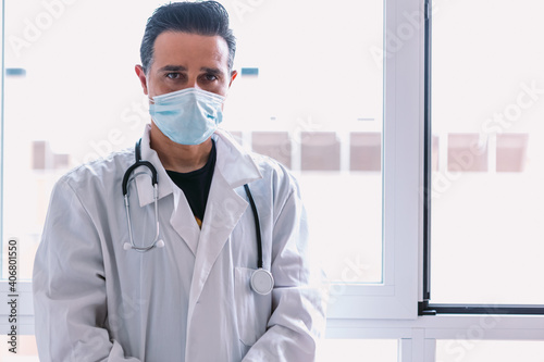 Black haired doctor wearing surgical mask, with white coat and stethoscope looking at camera, at a window. Medicine concept