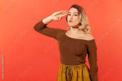 Portrait of responsible serious woman with blonde hair in brown blouse saluting commander, listening order with obedient expression. Indoor studio shot isolated on red background © khosrork