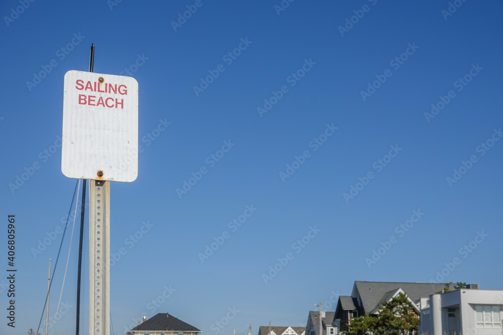 Fototapeta premium White sign on a metal post that says sailing beach against a clear cloudless blue sky.