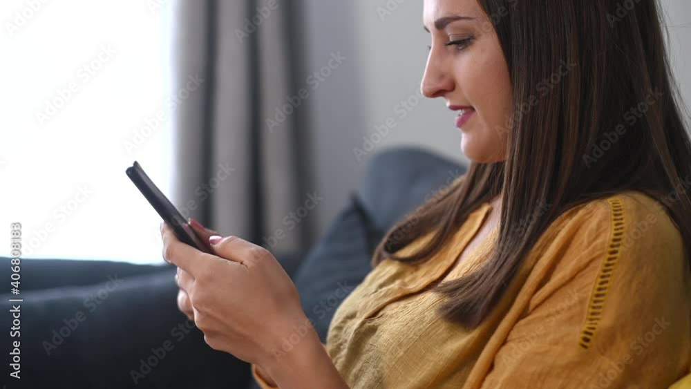 Side view a calm woman relaxing on comfortable couch, holding smartphone in hands, chatting in social networks, texting messages and scrolling news feed. A girl spends a leisure time with mobile phone Side view a calm woman relaxing on comfortable couch, holding smartphone in hands, chatting in social networks, texting messages and scrolling news feed. A girl spends a leisure time with mobile phone