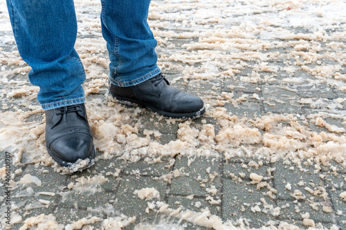 A man stands on the sidewalk in snow and mud. Black shoes and blue jeans close-up on the background of dirty snow. Ice on the road and sidewalk, the use of reagents, sand and salt.