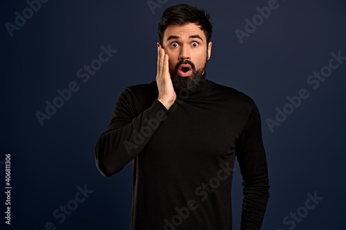 Shocked, speechless young handsome man with beard, gasping drop jaw and stare camera impressed, heard shocking rumor standing impressed Blue background, touch cheek after his crush kissed him