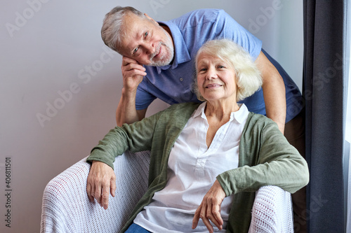 senior wife and husband posing at camera smiling on cozy couch at home, woman sitting while her husband stand behind her. portrait