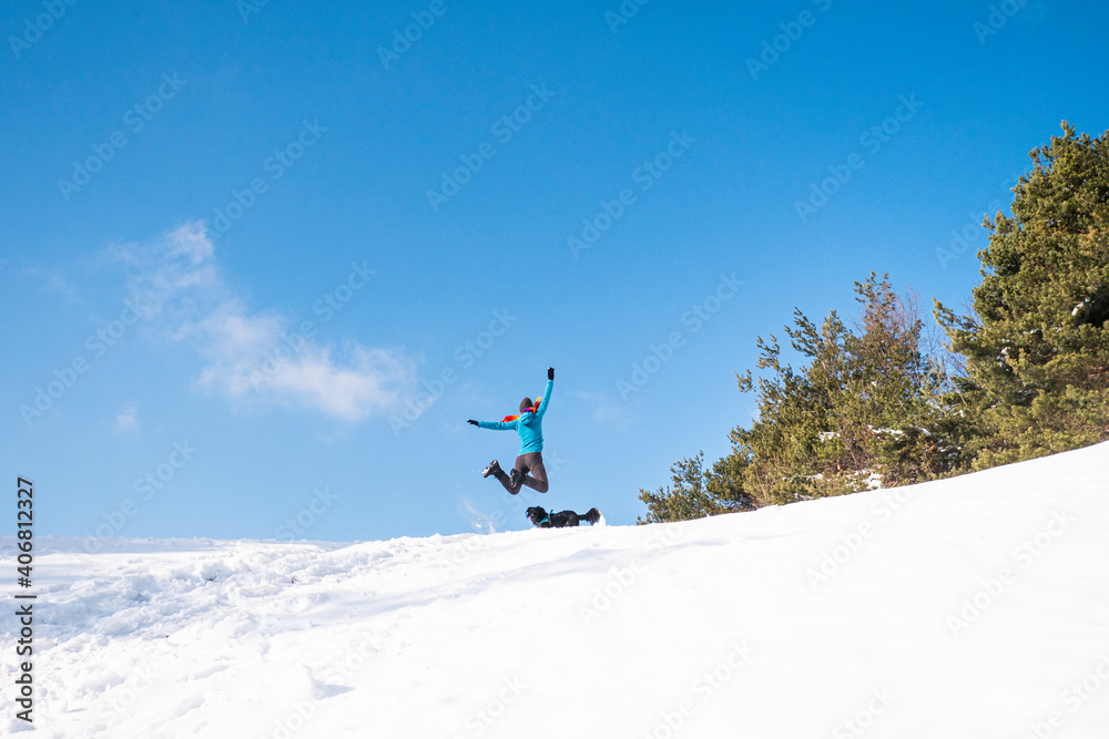 Young energetic female hiker jumping in the air on a sunny winter day. Winter hike in the mountains with a dog.