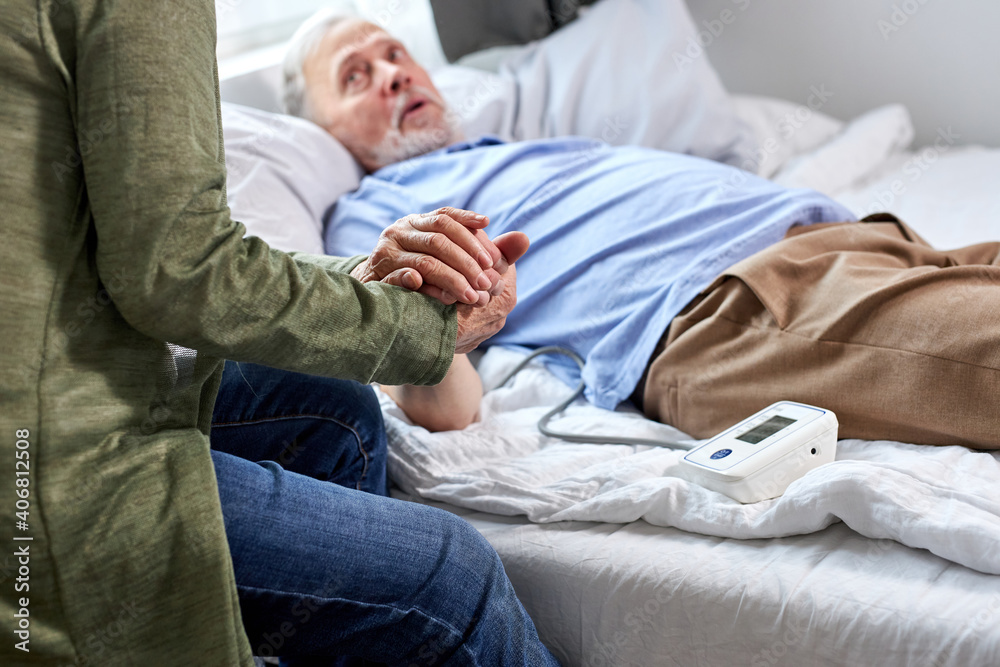 close-up photo of senior couple hands holding together, woman supports her sick husband lying on bed in hospital