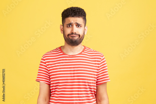 Frustrated unhappy man with beard in striped t-shirt looking at camera with sad dissatisfied expression, bad mood. Indoor studio shot isolated on yellow background