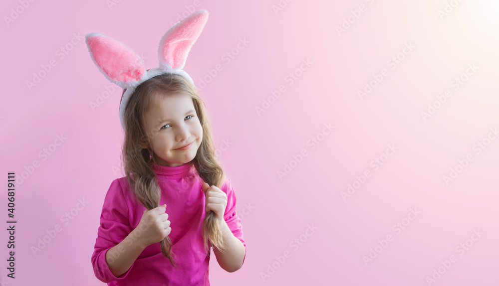 Happy Easter. A cute little girl in a pink sweater with bunny ears smiles cutely and holds herself by her ponytails of hair on a pink background. The child smiles and looks into the frame. Copyspace