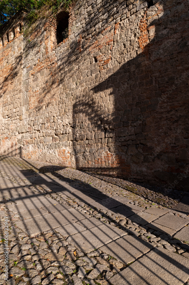 Obraz premium Detail of the shadow of a gate and of a woman with umbrella on the beautiful stone road in the medieval village of Monselice. Padua, Veneto.Italy.