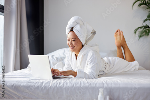 asian woman lying on bed at home and working on her laptop computer, wearing towel and bathrobe, in the morning. working from home, quarantine coronavirus concept