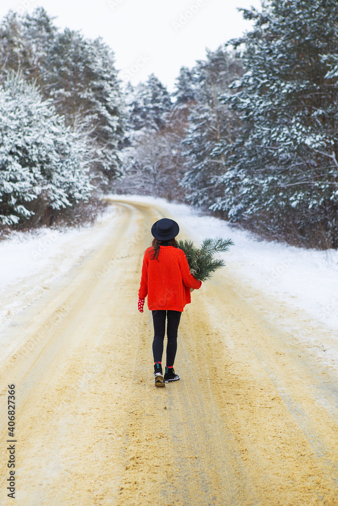 A girl in a red sweater and hat stands in the middle of a snowy road in a forest with pine branches. Weekend trip.