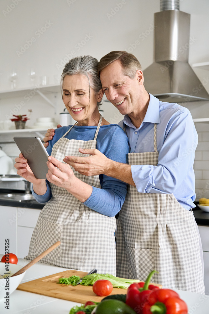 Smiling older senior 60s vegan couple preparing fresh vegetable salad in kitchen using tablet ordering food in grocery e shop or supermarket doing ecommerce shopping online cooking together at home. Smiling older senior 60s vegan couple preparing fresh vegetable salad in kitchen using tablet ordering food in grocery e shop or supermarket doing ecommerce shopping online cooking together at home.