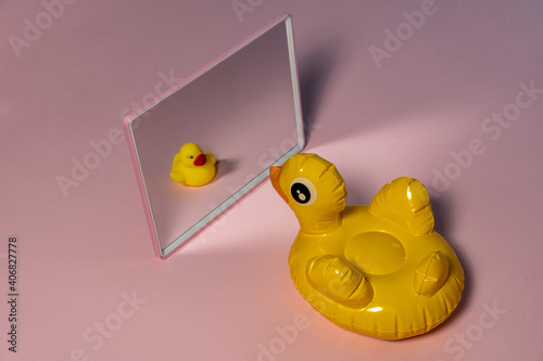 Rubber yellow duck in a mirror with a tiny reflection of itself.  Pink pastel background