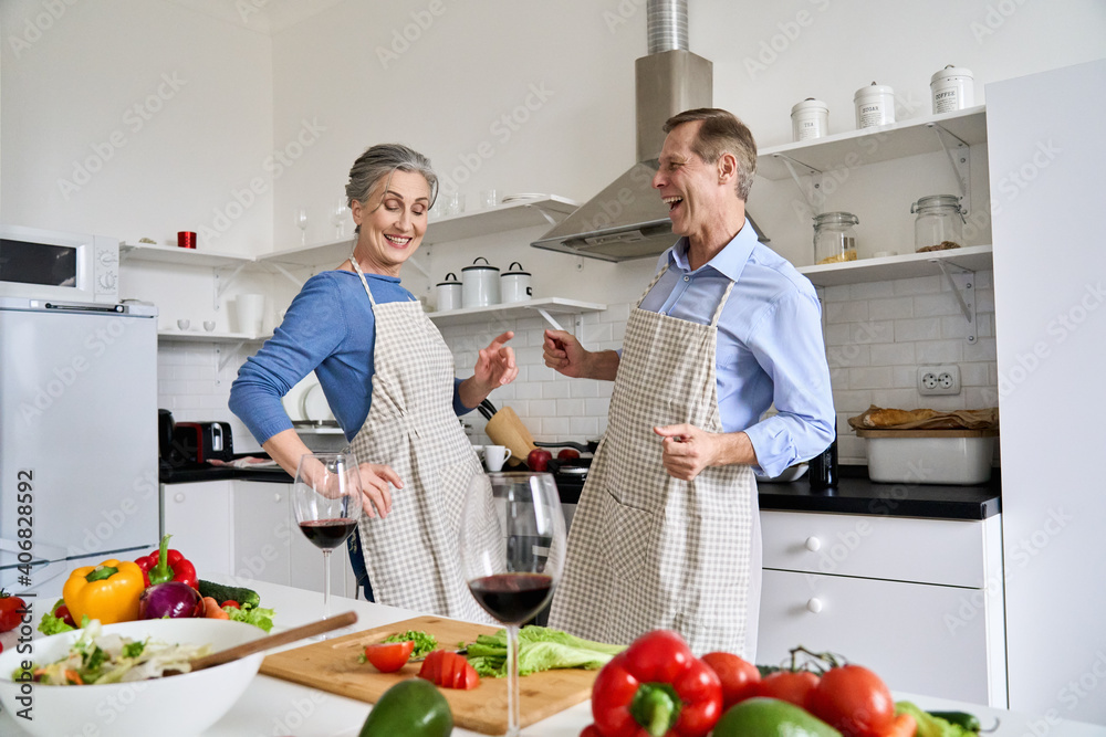 Happy active old senior mid aged 60s family couple enjoying dancing and cooking healthy meal together at home, having fun preparing romantic dinner, celebrating Valentines Day with wine in kitchen.
