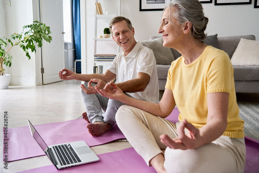 Obraz premium Happy healthy senior older 60s couple having fun learning to meditate, watching live online tv yoga class tutorial on laptop computer at home doing virtual training fitness workout sport exercises.