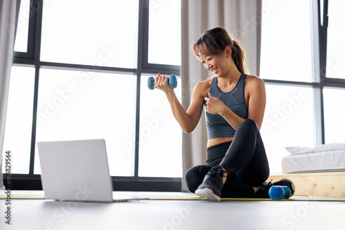 asian woman excercising at home, watching video tutorial on laptop, workout with dumbbells on the floor. sport online concept