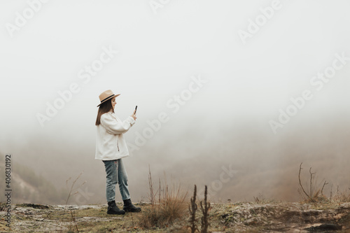 Girl standing on the peak edge and enjoying mountains view valley during heavy mist. She take the photo on smartphone.