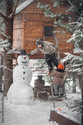 Cute boy and his little sister finishing making a snow man under the tree-house during the snowfall in winter in Russia. The put on a basket on it as a hat. Image with selective focus and toning