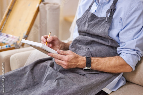 painter male is drawing on small canvas with pencil, making sketch, sitting on sofa in apron. cropped