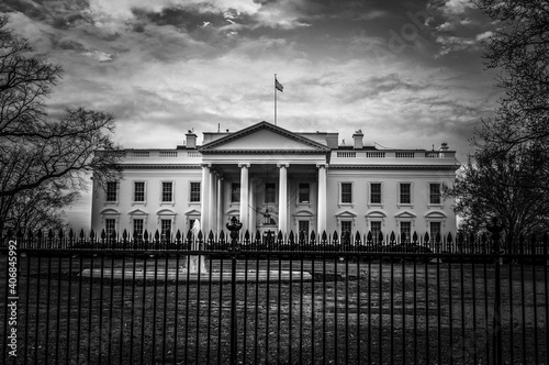 Dark black and white view of Whitehouse presidential residence in Washington DC with cloudy sky in background and fountain, grass and iron fence in the foreground. photo