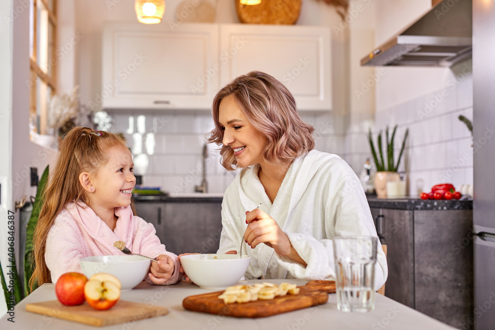 mother and daughter eating fruit and porridge. healthy nutrition for children, morning meal.caucasian family having breakfast in a light modern kitchen