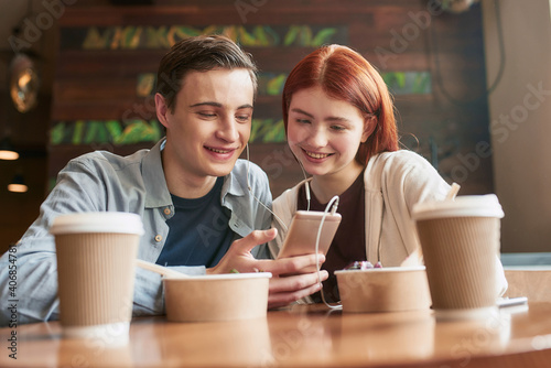 Happy teenage couple looking at the screen while watching something using smartphone and same pair of earphones, sitting in a cafe together on a daytime
