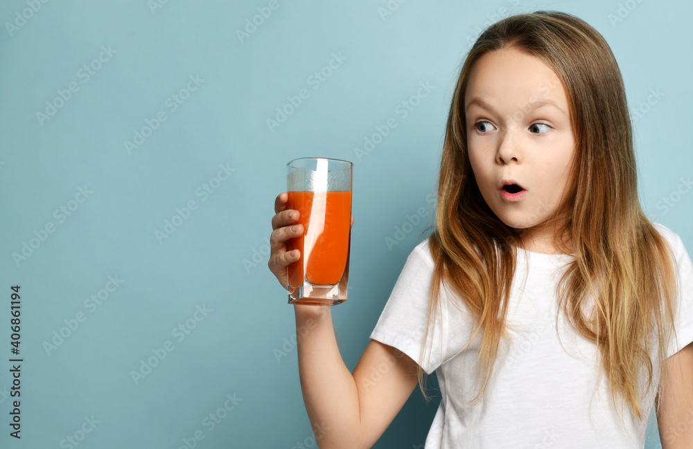 Little girl child kid drinking carrot orange juice surprised in white copy space blank t-shirt on light blue background. Health and diet concept. Healthy drink breakfast and clean food concept Little girl child kid drinking carrot orange juice surprised in white copy space blank t-shirt on light blue background. Health and diet concept. Healthy drink breakfast and clean food concept