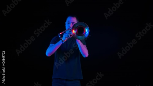 Front view of a musician playing the trombone in the studio against a black background in neon light. The thrombist performs music in the style of ska punk on a wind instrument. Close up. Slow motion. photo