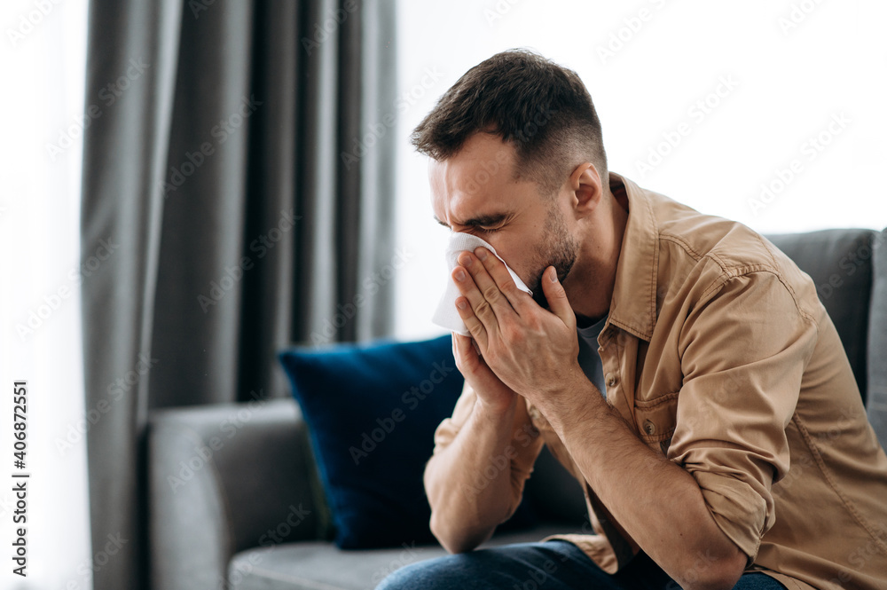 Unhealthy young man. Caucasian guy is sick with flu, he sneezes and blows his nose in a napkin while sitting on the couch at home Unhealthy young man. Caucasian guy is sick with flu, he sneezes and blows his nose in a napkin while sitting on the couch at home