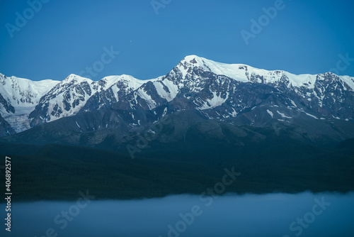 Atmospheric mountains landscape with dense fog and great snow mountain top under twilight sky. Alpine scenery with big snowy mountains over thick fog in night. High snow pinnacle above clouds in dusk.