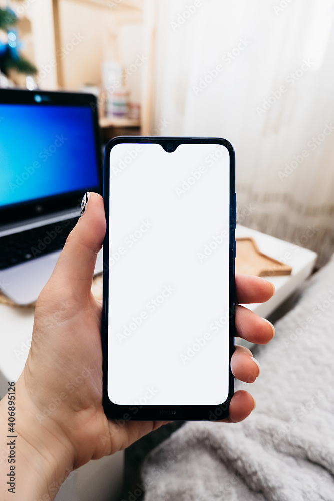 smartphone with white screen in hand on background of workplace in minimalist scandi style with laptop, space for text, vertical content, selective focus smartphone with white screen in hand on background of workplace in minimalist scandi style with laptop, space for text, vertical content, selective focus