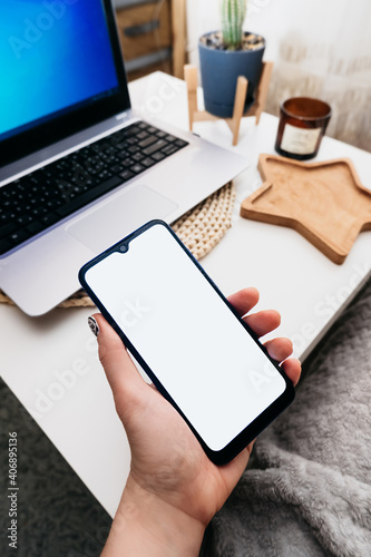 smartphone with white screen in hand on the background of the workplace in a minimalist scandi style with a laptop, cactus and candles, space for text, vertical content, selective focus